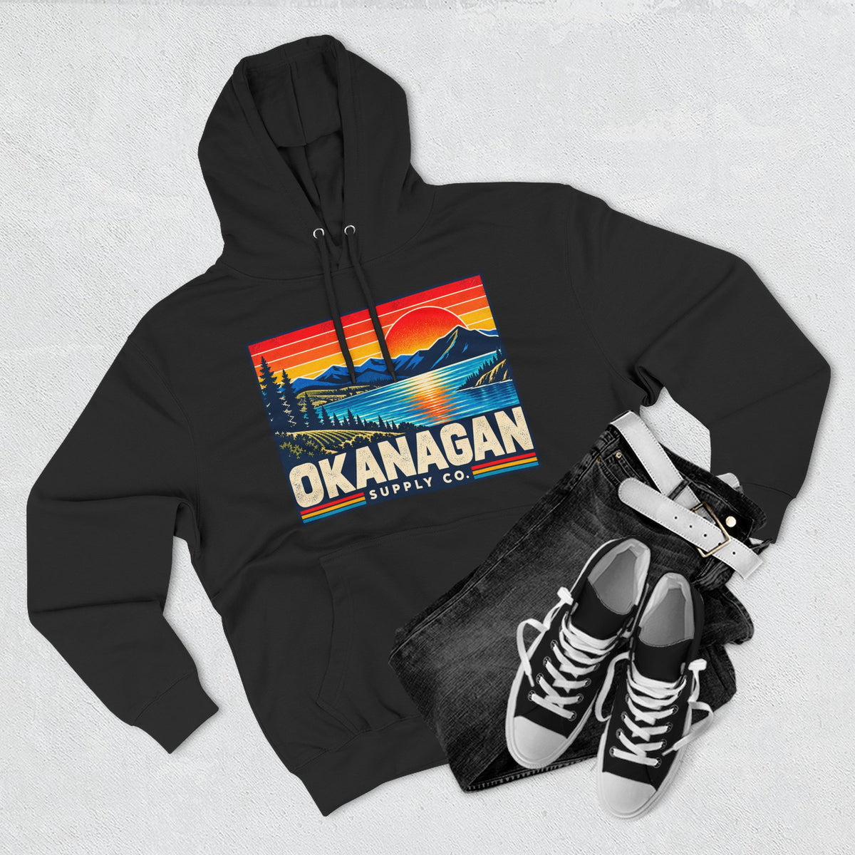 Okanagan Supply Co. Retro Sunset Hoodie in black, flat lay with jeans and sneakers