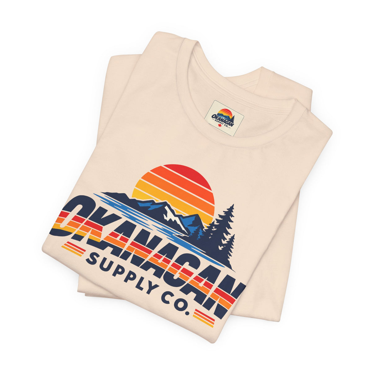Okanagan Supply Co. Vintage Outdoors Tee in soft cream, folded to show the retro sunset, mountain and pine print and inside collar label